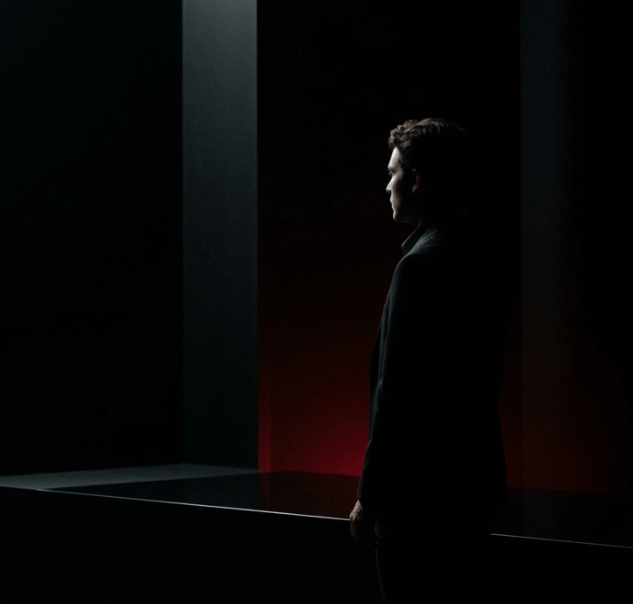 Fotografia dramática e escura em silhueta de um homem de perfil em terno escuro, virado para a direita, em pé entre duas grandes colunas escuras. Um brilho vermelho neon vertical brilha por trás das colunas, delineando sutilmente a figura.